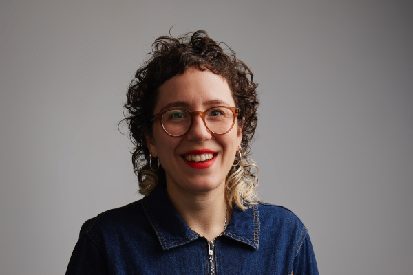 Portrait of a white woman in her 30s looking at the camera. She's wearing a black shirt, has curly hair with bleached ends and a short fringe, wears bright red lipstick, light brown glasses, and a gold watch. She has a small tattoo on her wrist.