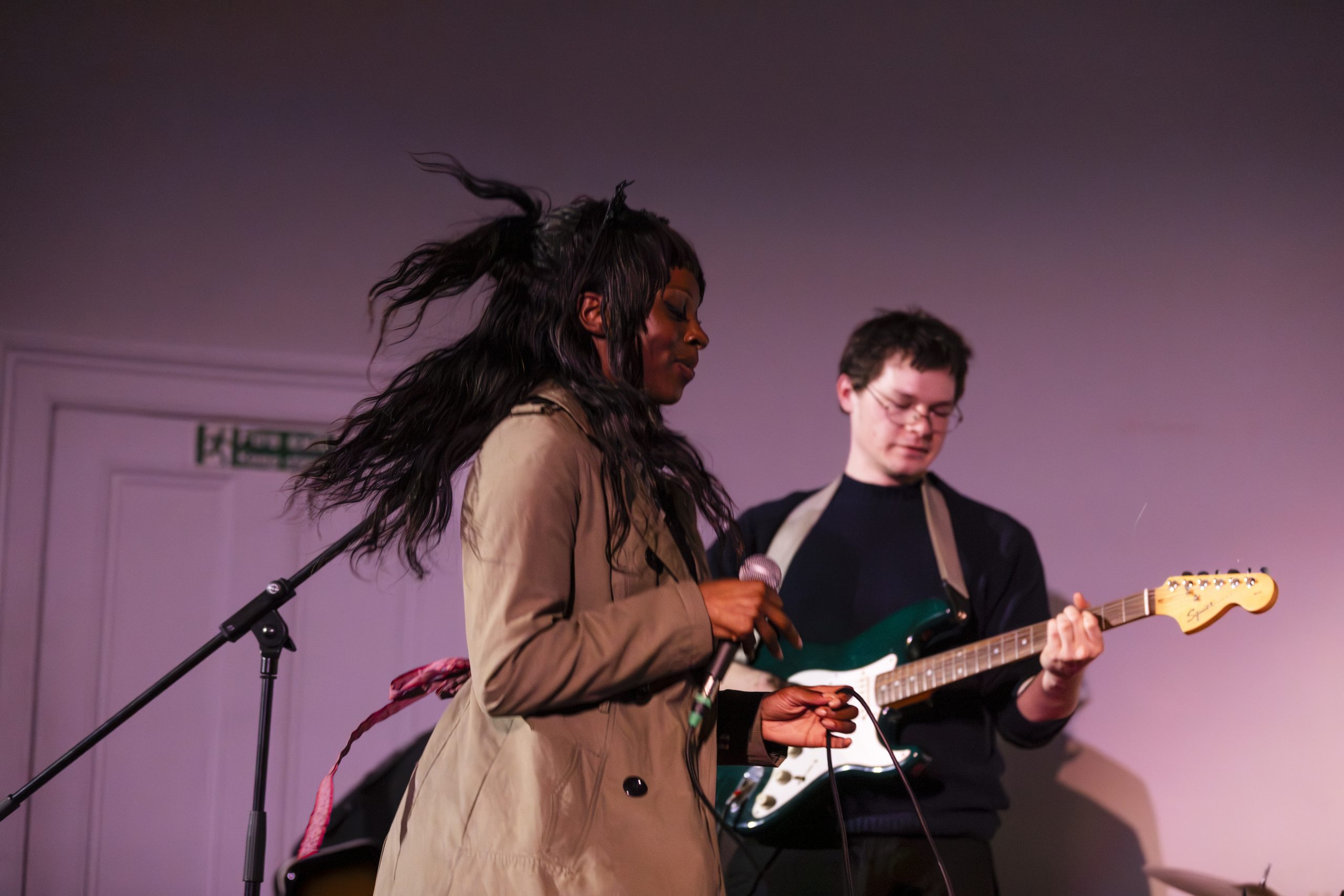 A young black woman is dancing in a beige trench coat, behind is a young white man playing guitar.