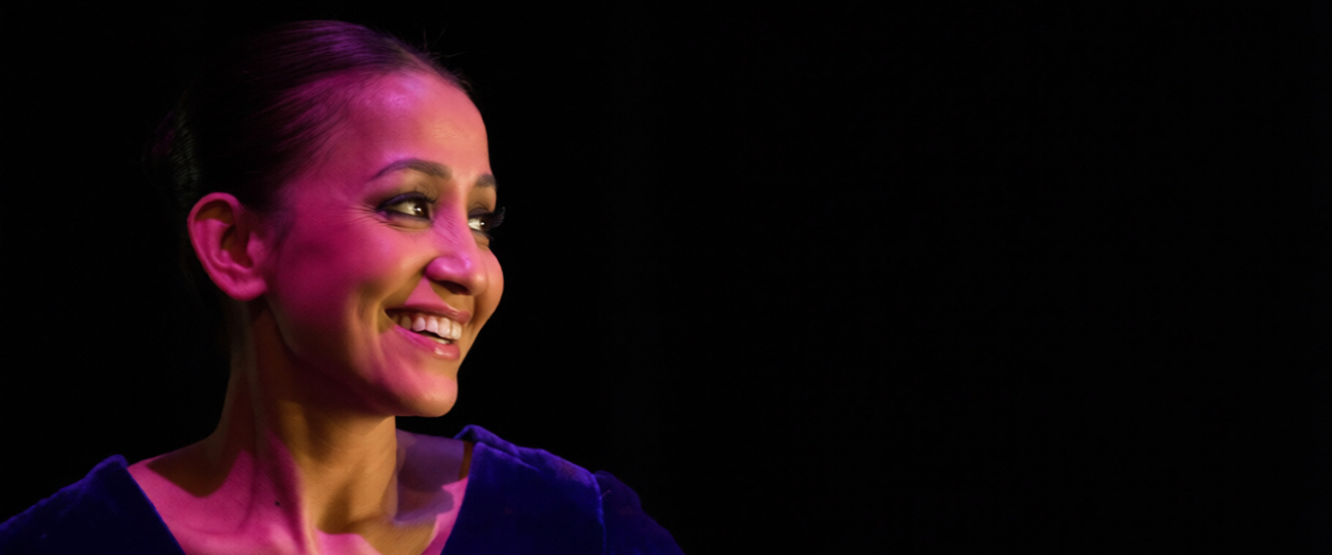 A smiling woman wearing a blue velvet top, illuminated by pink and orange stage lighting against a black background