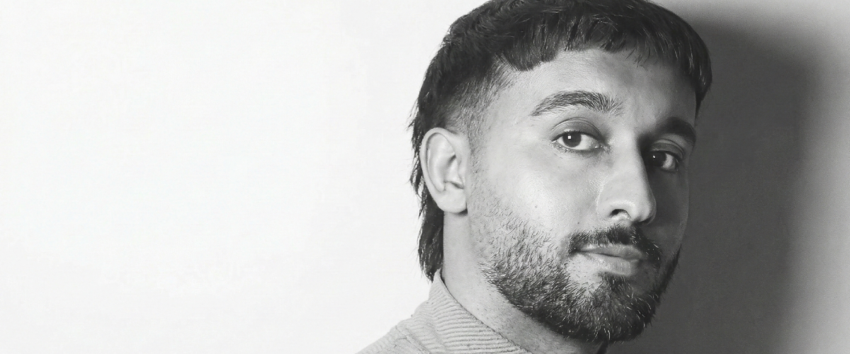 A black and white portrait of a man in a three-quarter pose, looking directly at the camera, wearing a textured zip-up sweater against a plain background.