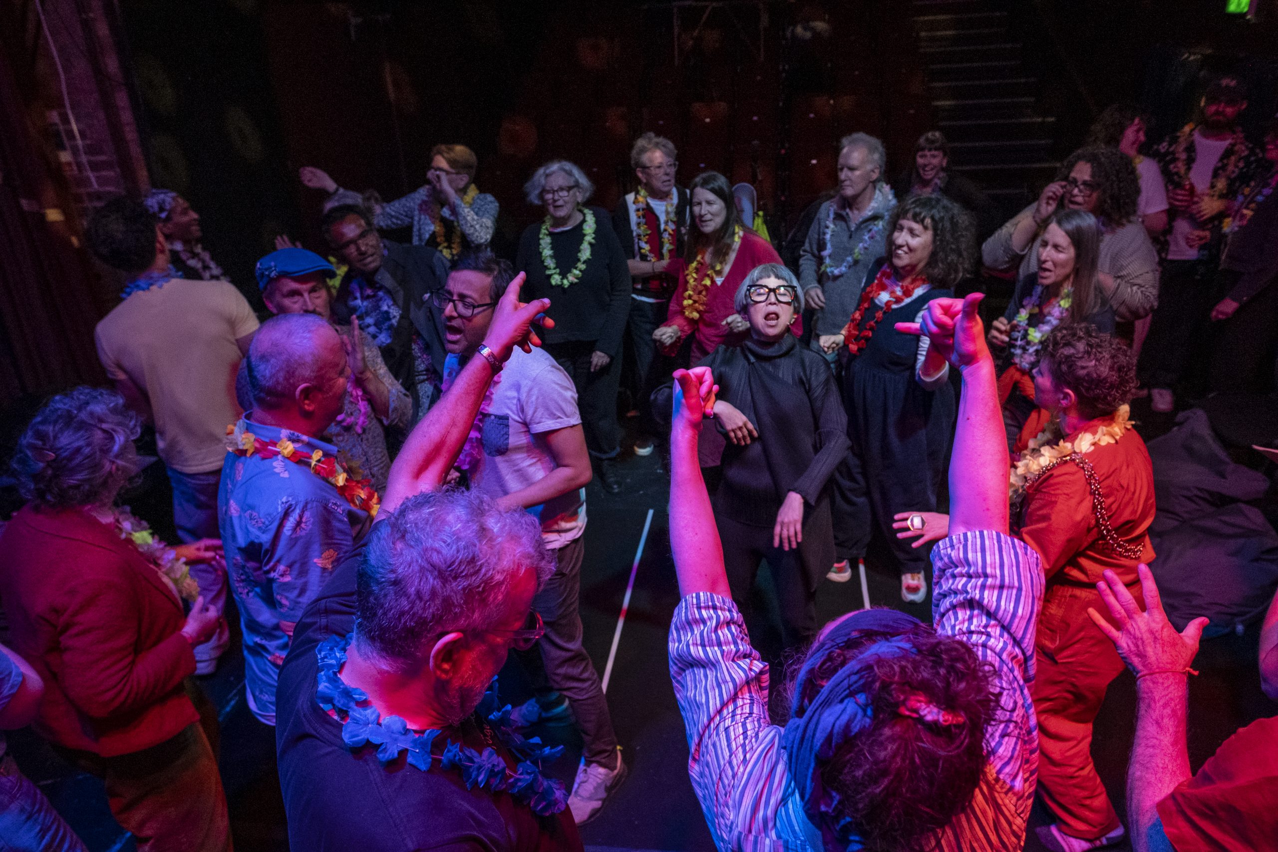 A diverse group of people dance and gesture enthusiastically in a dimly lit theater space, many wearing colorful leis. The scene feels energetic and communal, with raised arms and expressive movement under vibrant pink and blue stage lighting.
