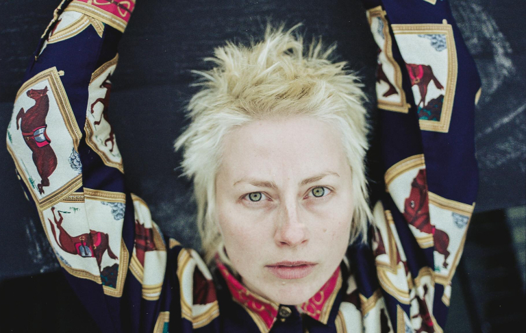A white person with short, bleached-blond hair looks directly at the camera with their arms raised overhead, wearing a patterned button-up shirt. The dark background and symmetrical pose give the portrait a dramatic, stylized feel.