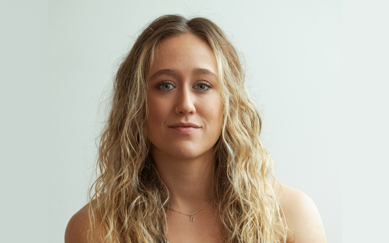 A woman with long, wavy blonde hair and blue eyes, wearing a simple necklace, photographed against a light, neutral background. She is facing the camera with a calm, neutral expression.