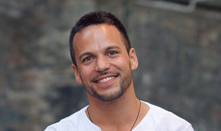 A smiling man with short dark hair and light facial hair, wearing a white shirt, photographed outdoors against a blurred stone background. He is looking directly at the camera.