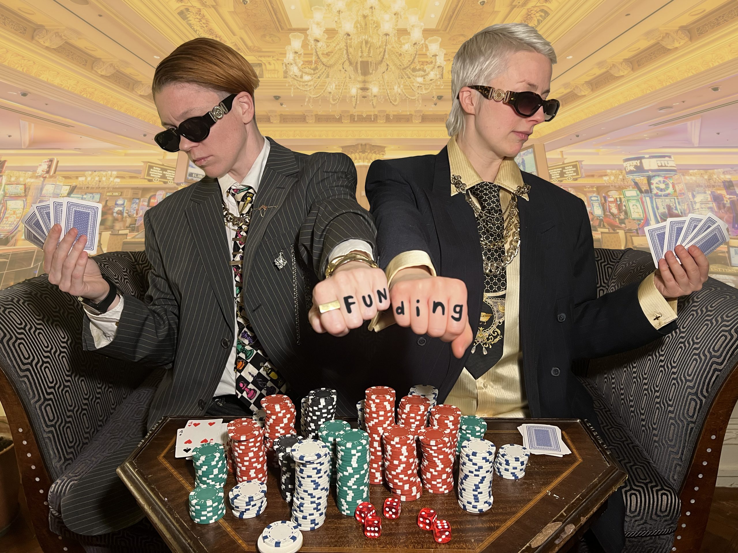 Two stylish white people in suits and sunglasses sit back-to-back at a casino table piled with poker chips, cards, and dice. Their fists meet in the foreground, with letters on their knuckles spelling “FUNding,” set against a luxurious casino interior.