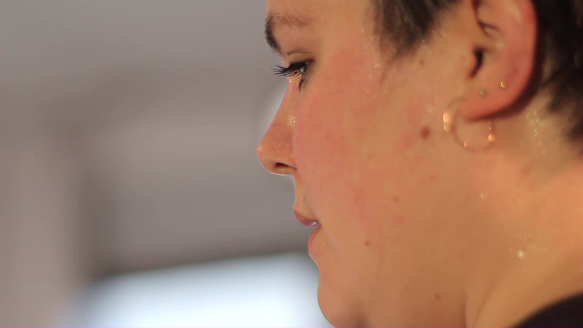 Slightly blurred close-up profile of a person with short hair and earrings, sweat on their face, looking focused.