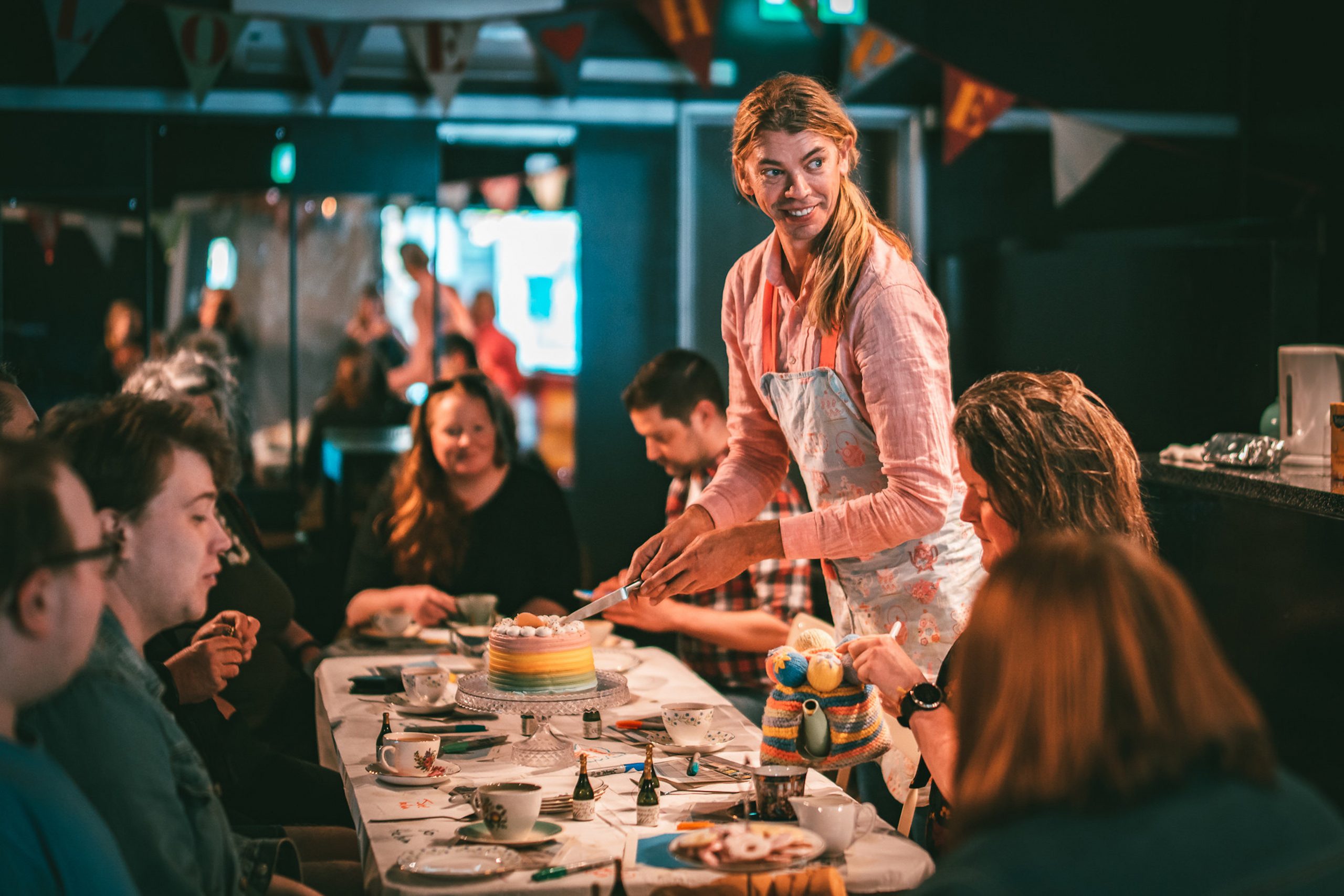 A tall, smiling person with shoulder length blond hair in a low pony-tail, wearing a floral apron serves a rainbow cake to a long table of smiling guests at a cosy tea party. The room is warmly lit, with festive bunting above.