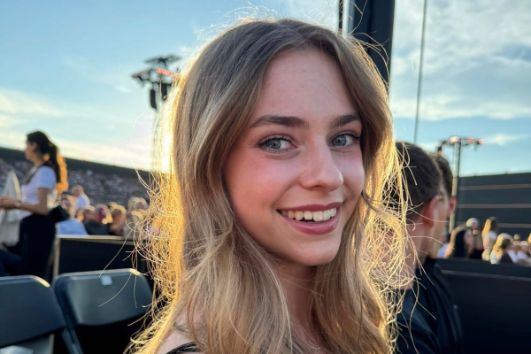 Smiling white young woman with long blonde hair sitting in an outdoor venue, wearing a black sleeveless top, with rows of seats and a crowd behind her under a clear sky.