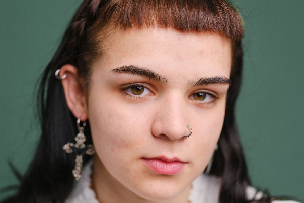 A close-up portrait of a white person with dark hair and short bangs, wearing a nose ring and earrings, looking directly at the camera against a green background. They are dressed in a light-colored top with ruffled details.