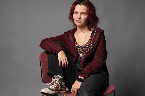 A seated studio portrait of a person with shoulder-length red hair, wearing a burgundy hoodie, patterned top, and black jeans. They sit casually on a chair against a gray backdrop, with one leg bent and an arm resting on their knee.