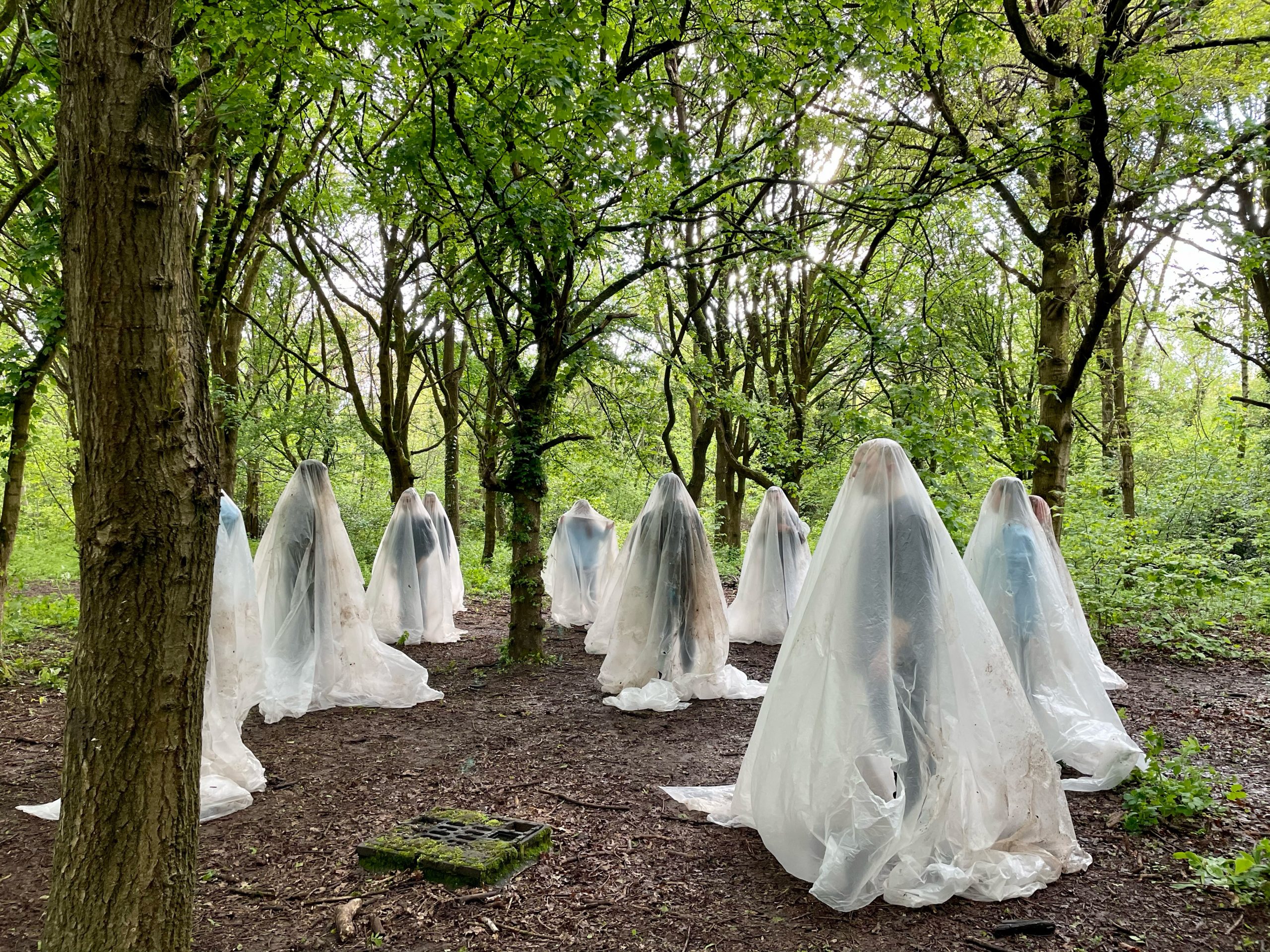 An outdoor performance featuring multiple human figures standing in a forest, each draped in translucent white plastic sheeting among the trees.