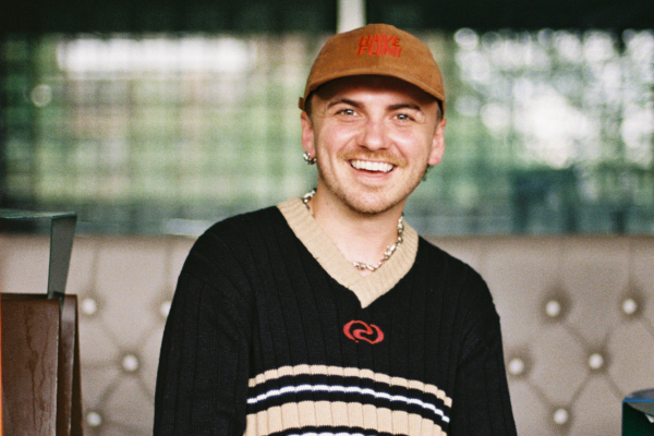Smiling white person wearing a brown baseball cap and a black sweater with a striped V-neck, seated indoors with a softly blurred background.