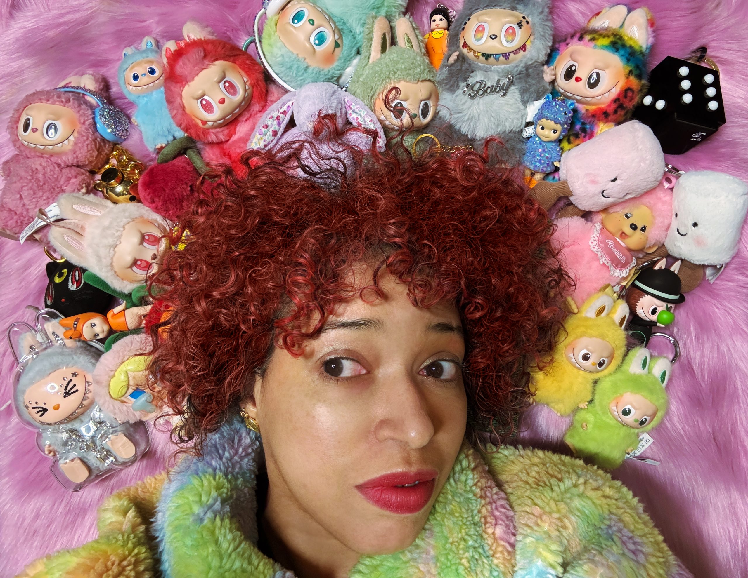 A mixed race woman in her forties with pink lipstick and red curly hair, lies on pink fake fur backdrop. She is wearing a multicoloured pastel fleece jacket. Her eyes look to the left as if in thought. Arranged around her hair is a colorful collection of cute trinkets and plush keychains packed tightly together, fuzzy pastel characters with big eyes and toothy smiles and, rhinestone-covered charms