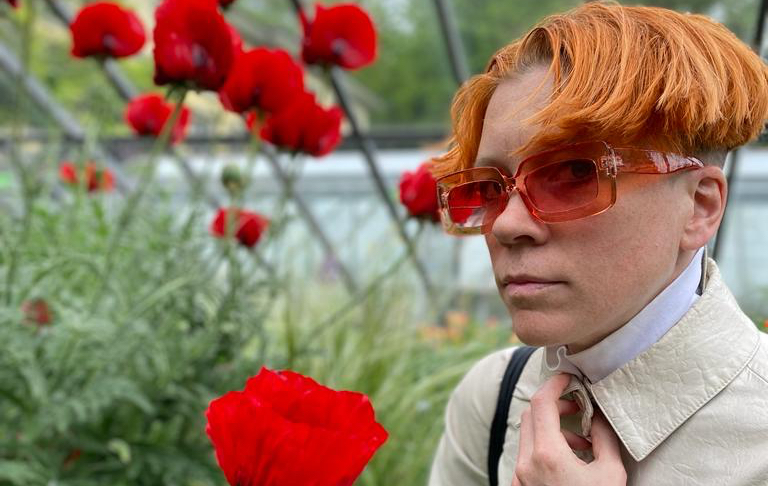 A white person with short orange hair and tinted glasses stands in a greenhouse among tall red poppies, adjusting the collar of a light-colored coat. Soft natural light and greenery frame the scene, creating a calm, contemplative mood.