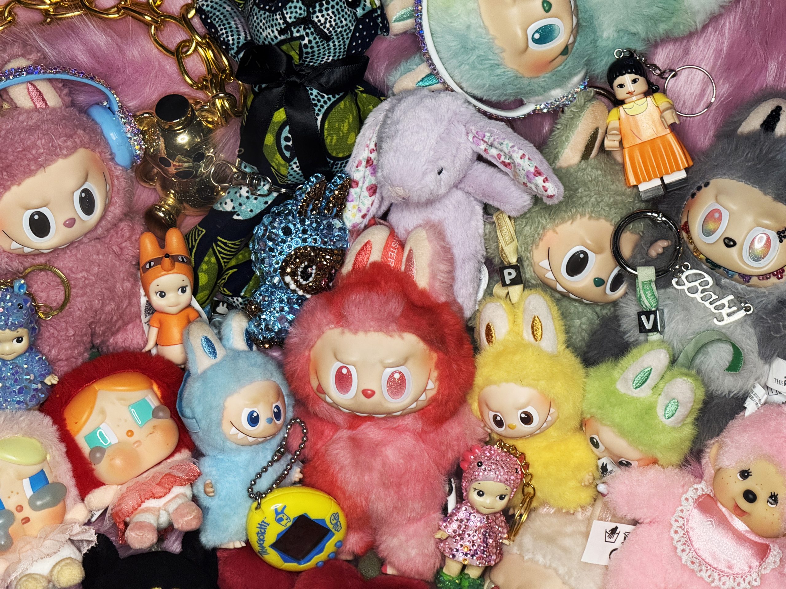 A colorful collection of cute trinkets and plush keychains packed tightly together, featuring labubus, fuzzy pastel characters with big eyes and toothy smiles in pink, red, blue, yellow, green, and purple, along with customised sonny angels, small doll figures, rhinestone-covered charms, and metal key rings, crying doll figures and tamagotchis digital pet keychains. creating a playful, collectible aesthetic.