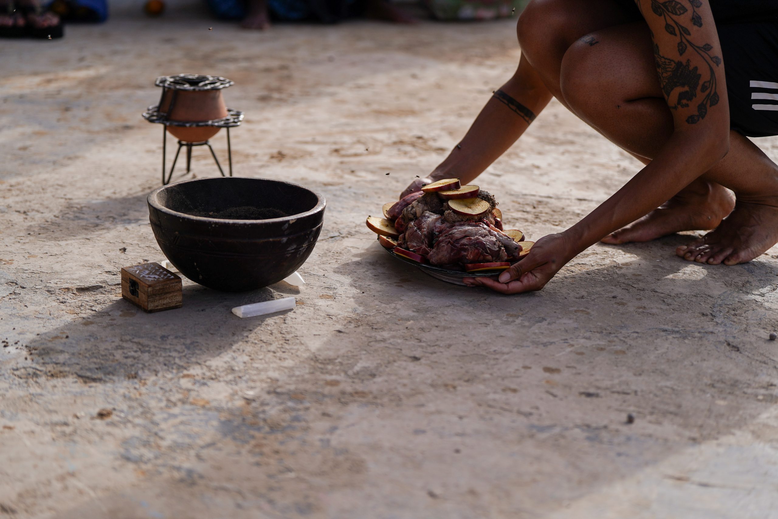 A brown skinned tattooed person with only torso, arms and legs visible, is placing down a plate of goat meat surrounded by sliced apples, onto a concrete ground. In front of the plate is a brown calabash encircled by oblong pieces of selenite, a white crystal. Directly behind the bowl is a small wooden box, and on the other side of the bowl sits a ceramic and iron incense burner.