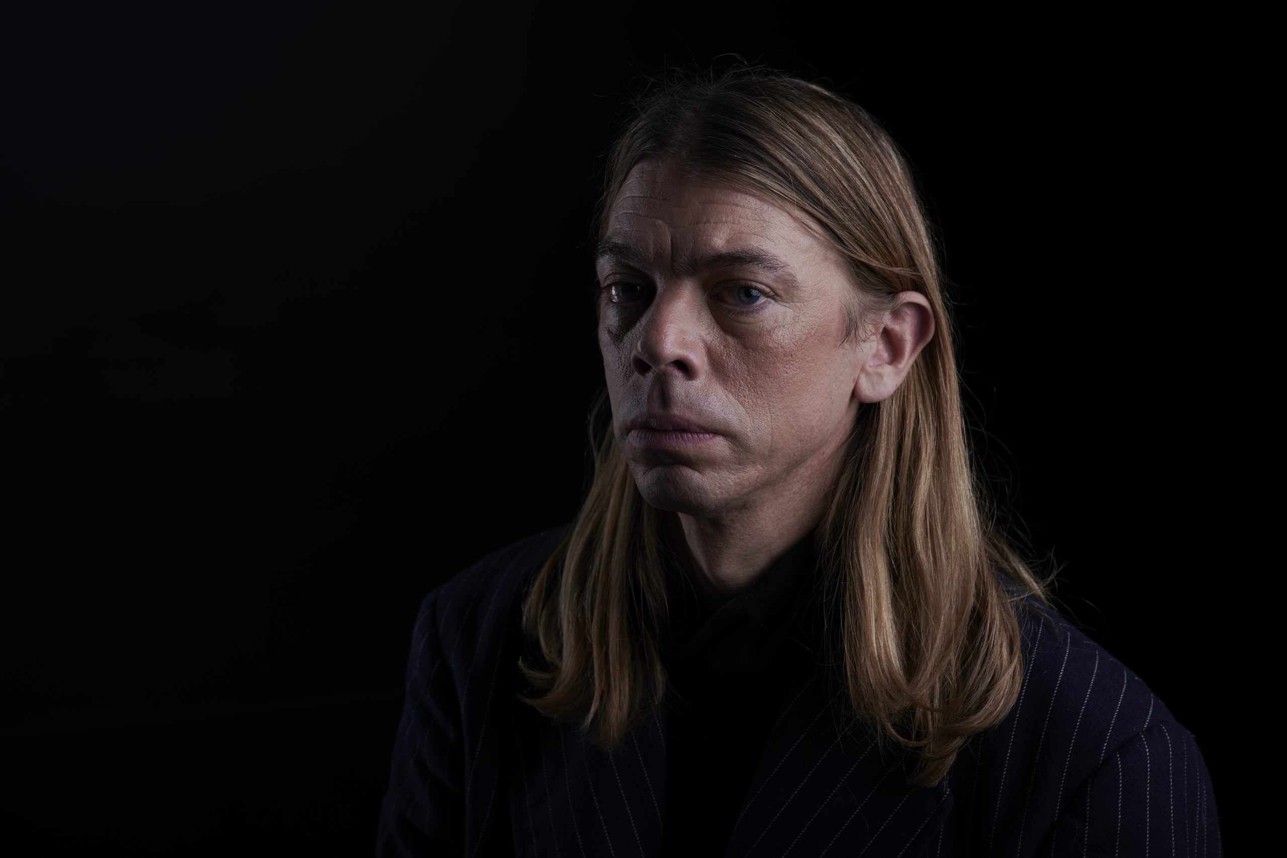 A studio portrait of a person with long, straight light-brown/blonde hair parted in the middle, wearing a dark jacket. They are shown from the chest up against a black background, lit from one side, with a serious, contemplative expression and their gaze directed slightly off-camera.