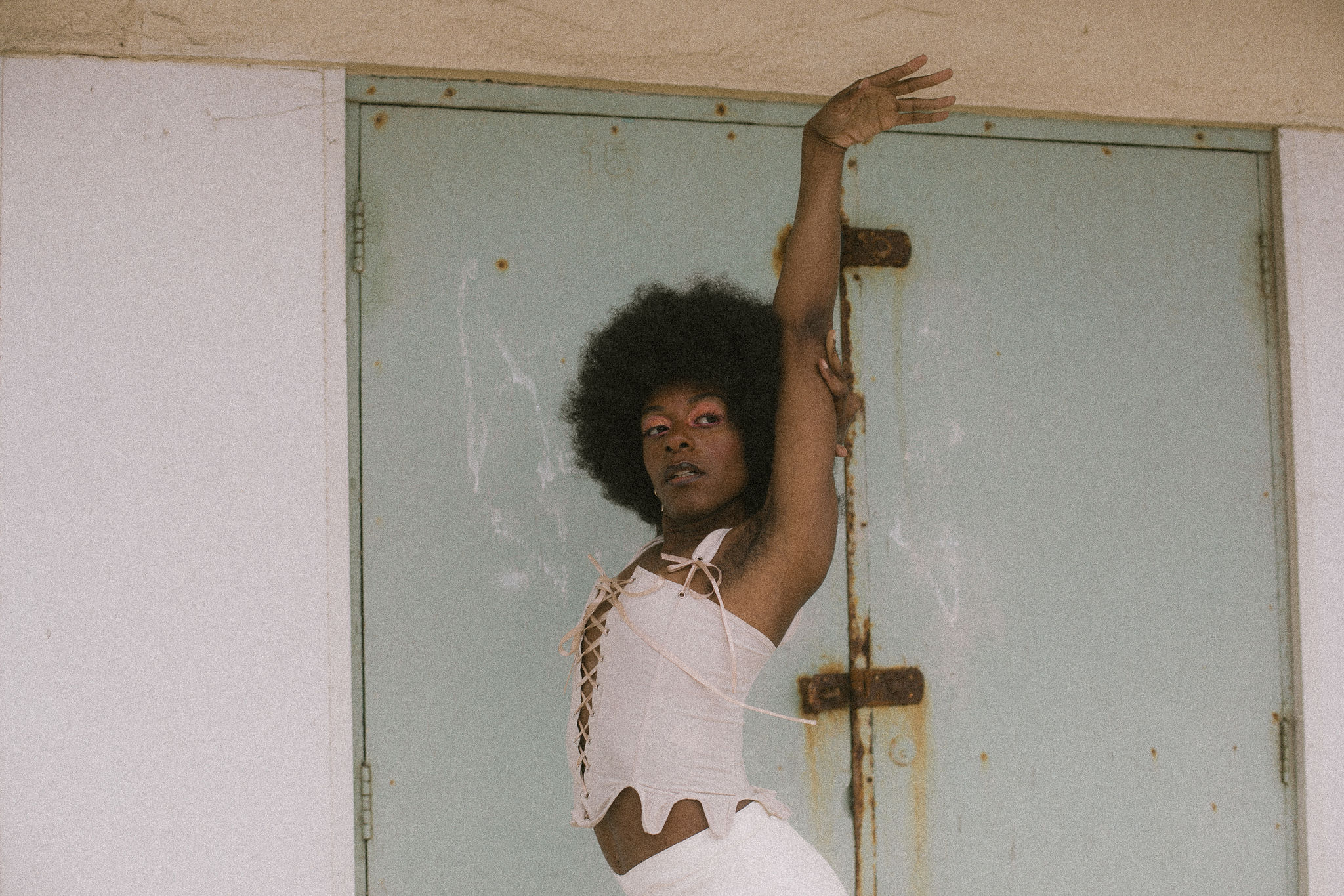 A slim black person with an afro stands in a white top to the side. They are facing the camera and have their hands elegantly above their head.