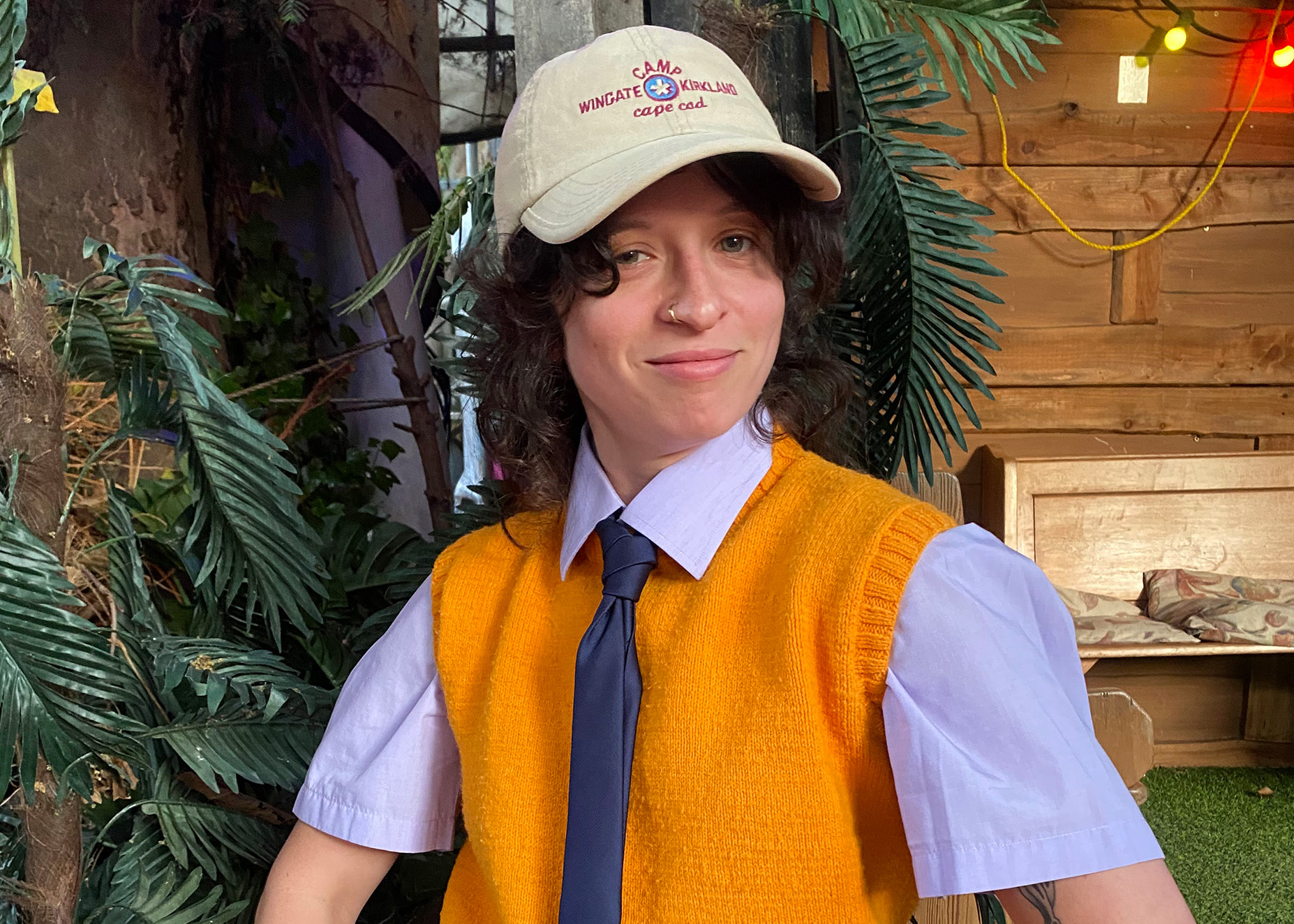 A slim Jewish person with curly dark hair and a nose ring sits smiling in front of wooden panelling and plants wearing a pale blue shirt, mustard yellow vest, dark blue tie and beige cap.