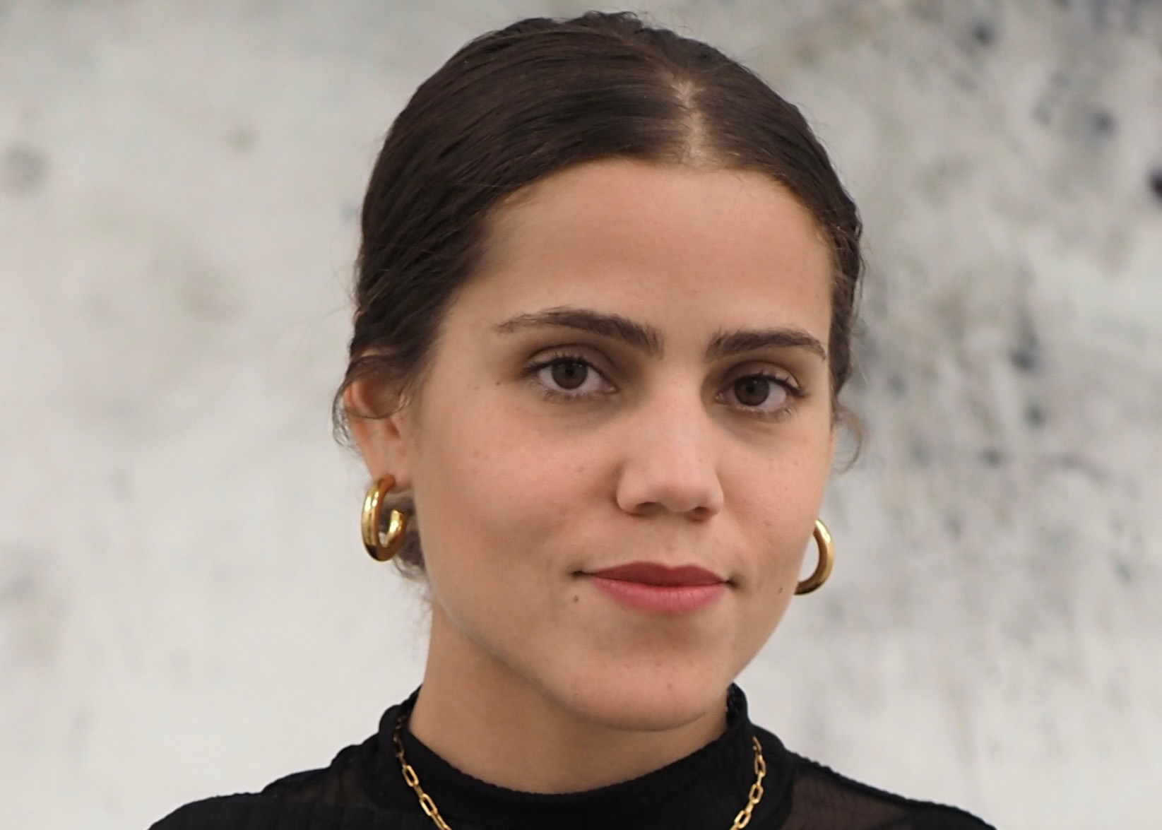 Gabriela has dark hair and is wearing a black high necked top with a fine chain necklace with a small pendant and hoop earrings.