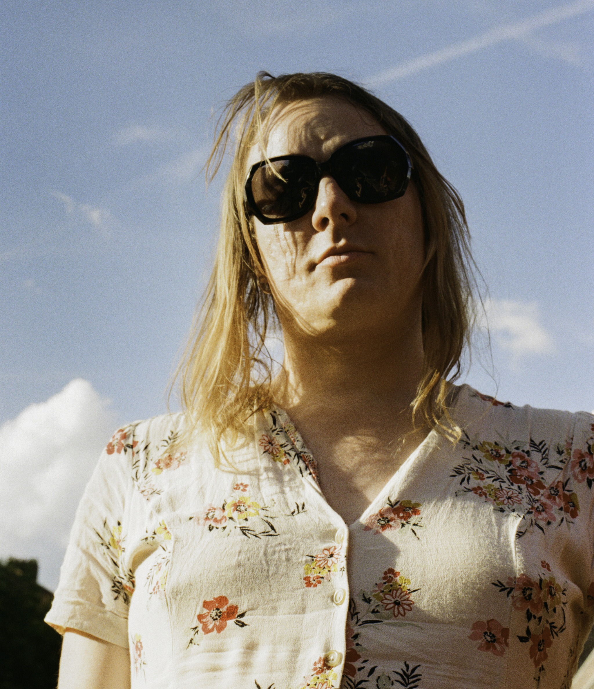 Carle is a slim white person with blonde hair. She is facing the camera with large black sunglasses and a pale ditsy floral dress.
