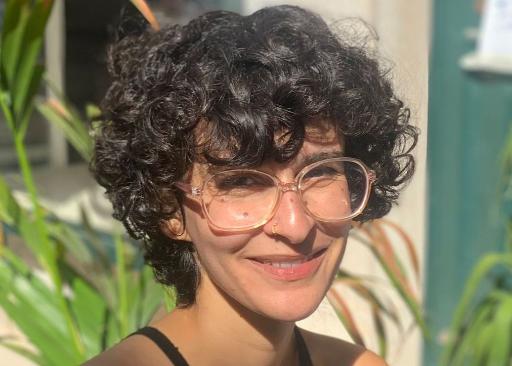 Miriam has short, dark curly hair and wears clear framed glasses. She is smiling, sat in front of a palm plant.