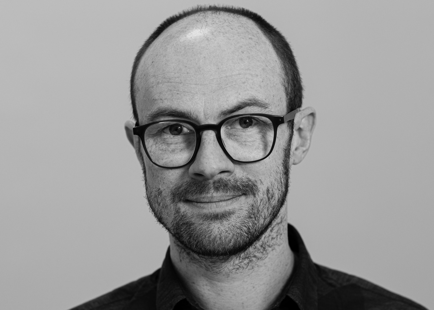 Jon is a white man with a shaved head, glasses and stubble. He is wearing a dark shirt.