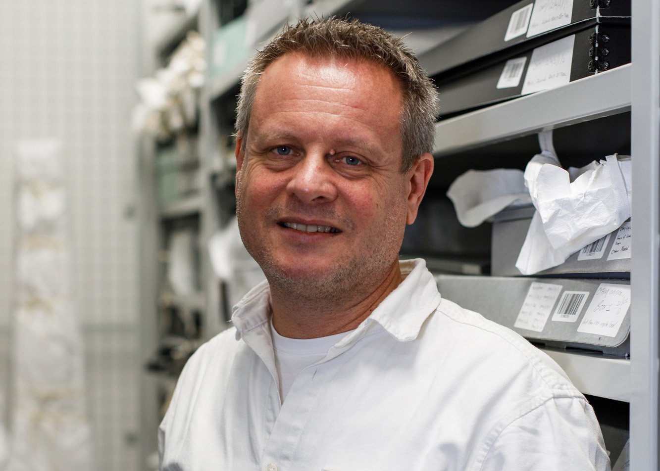 Julian is a white man with short salt and pepper hair. He is standing in an archive space, wearing a white shirt.