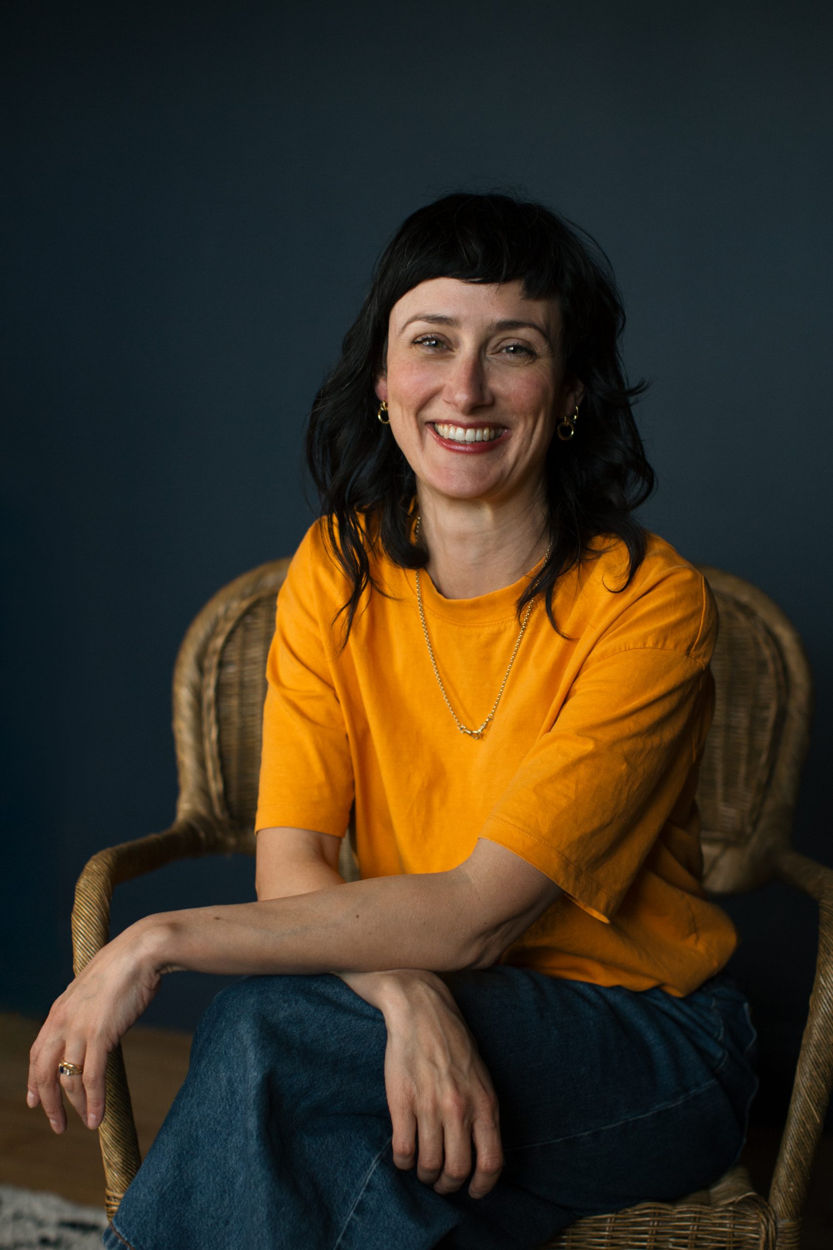 Sally is a slim, white, femme presenting person. She is sat, portrait style on a wicker chair, wearing a yellow t-shirt. Sally has long dark hair and is smiling brightly.