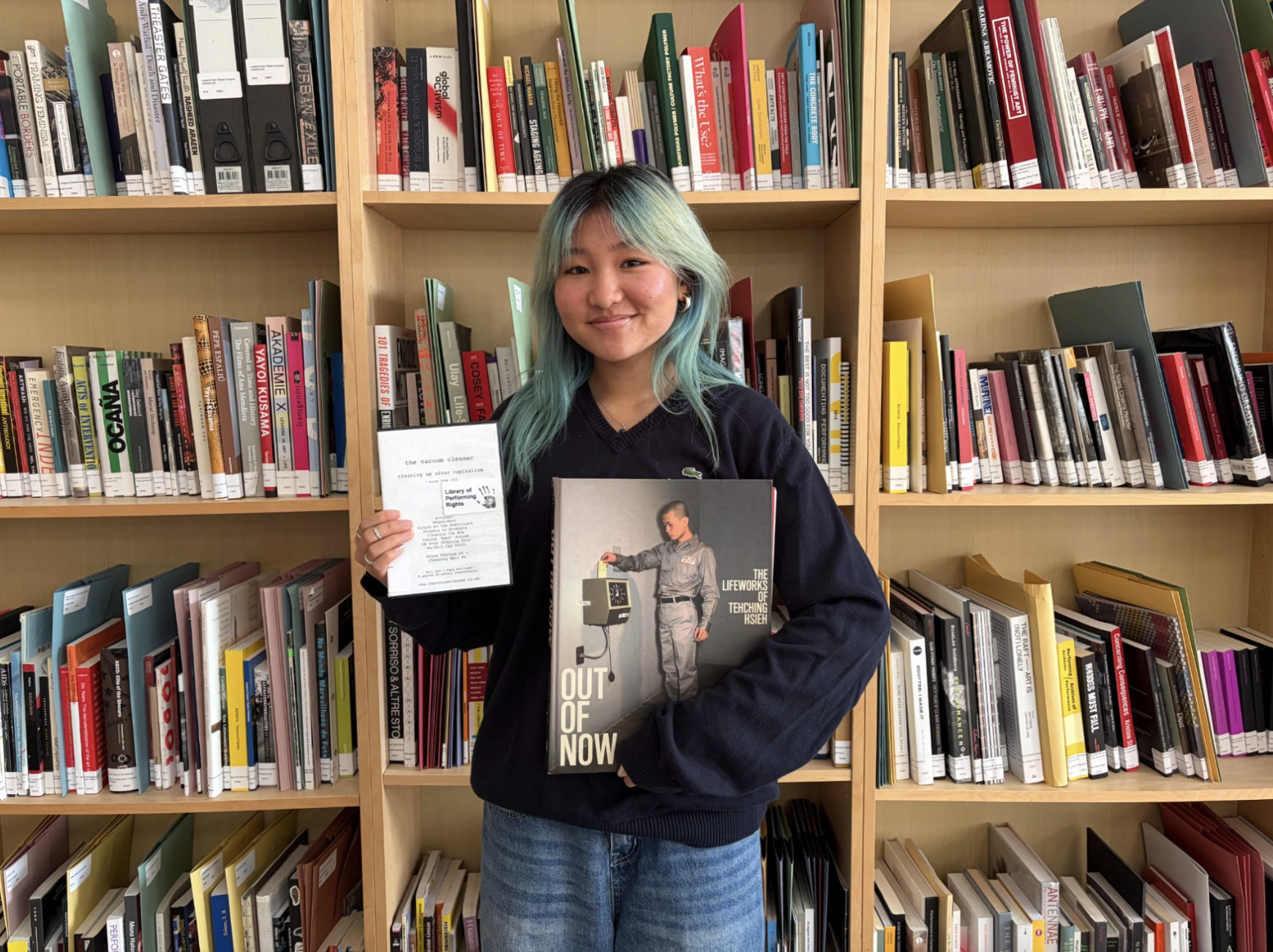 Kenn has long blue hair and wears a dark hoody with blue jeans. She is Asian-Canadian and is holding a DVD and a book.