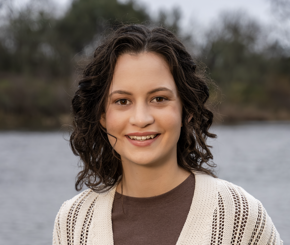 Olivia stands smiling at the camera. She has shoulder length dark curly hair and wears a brown top with a cream crochet cardigan.