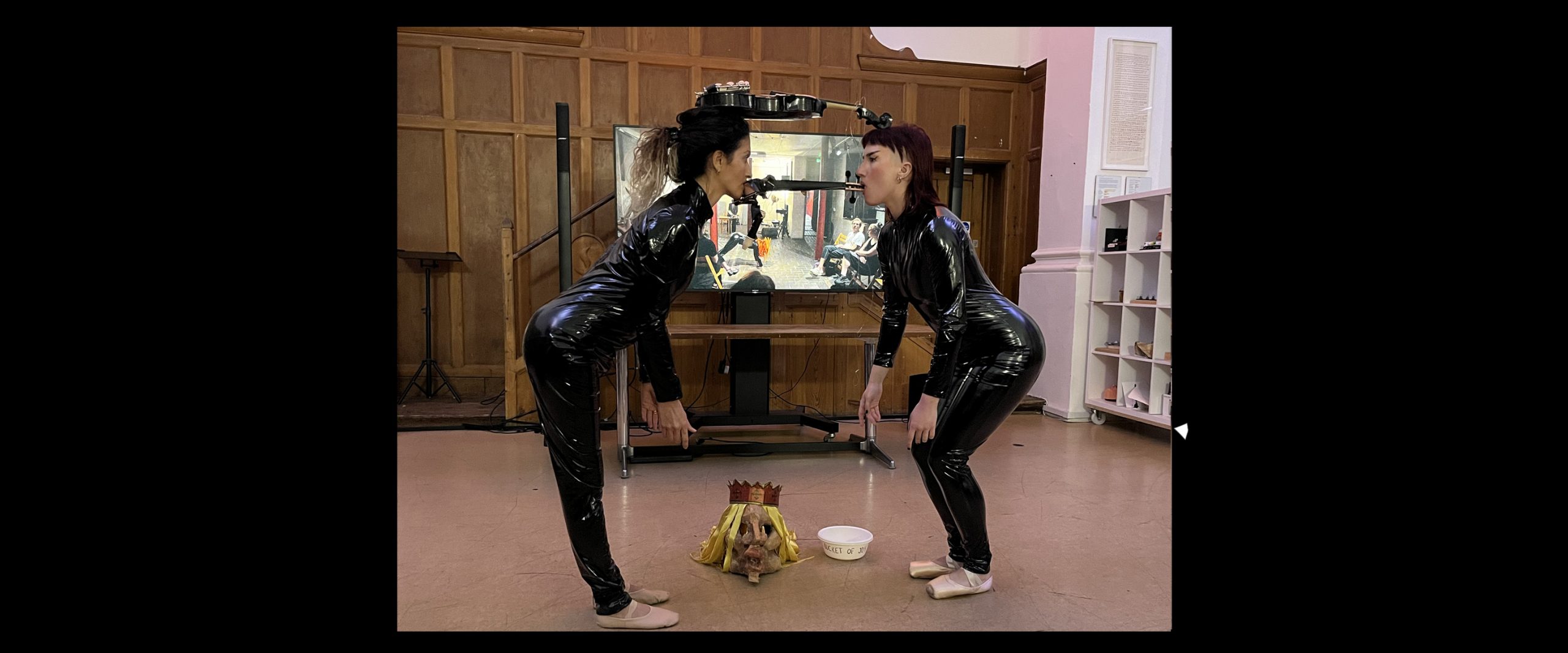Two people in black latex bodysuits stand facing each other. They are balancing a violin between them and hold wood between their teeth.