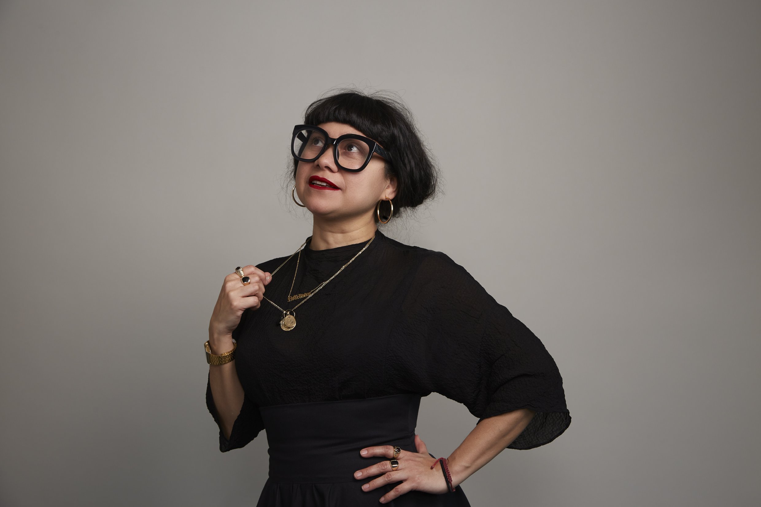 teresa has dark hair in a bob with a fringe, wears red lipstick and glasses and is holding a gold chain necklace.