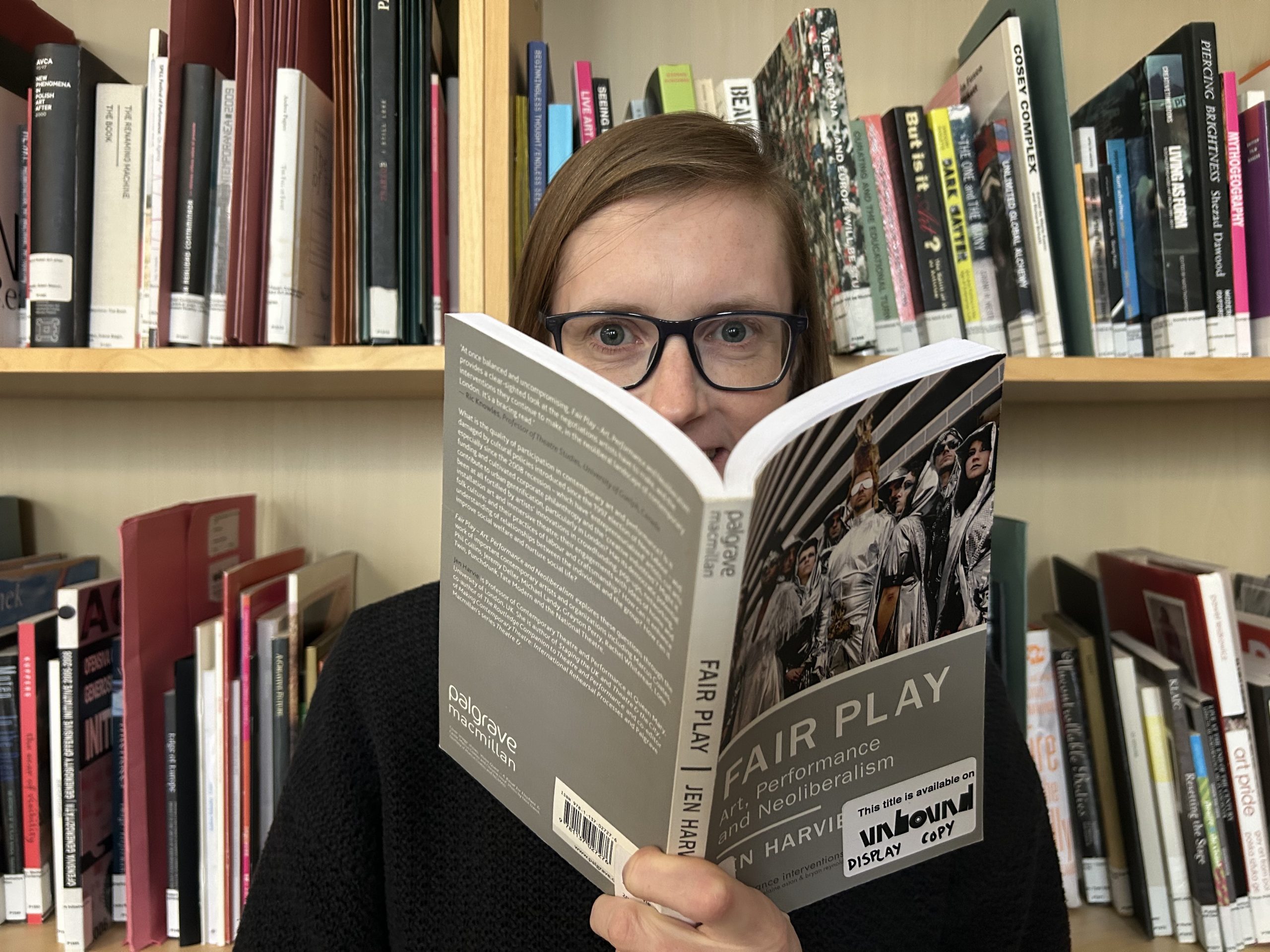 Charlotte is pale with sandy brown straight hair in a side parting. She wears dark square rimmed glasses and is reading from Fair Play - Jen Harvie.