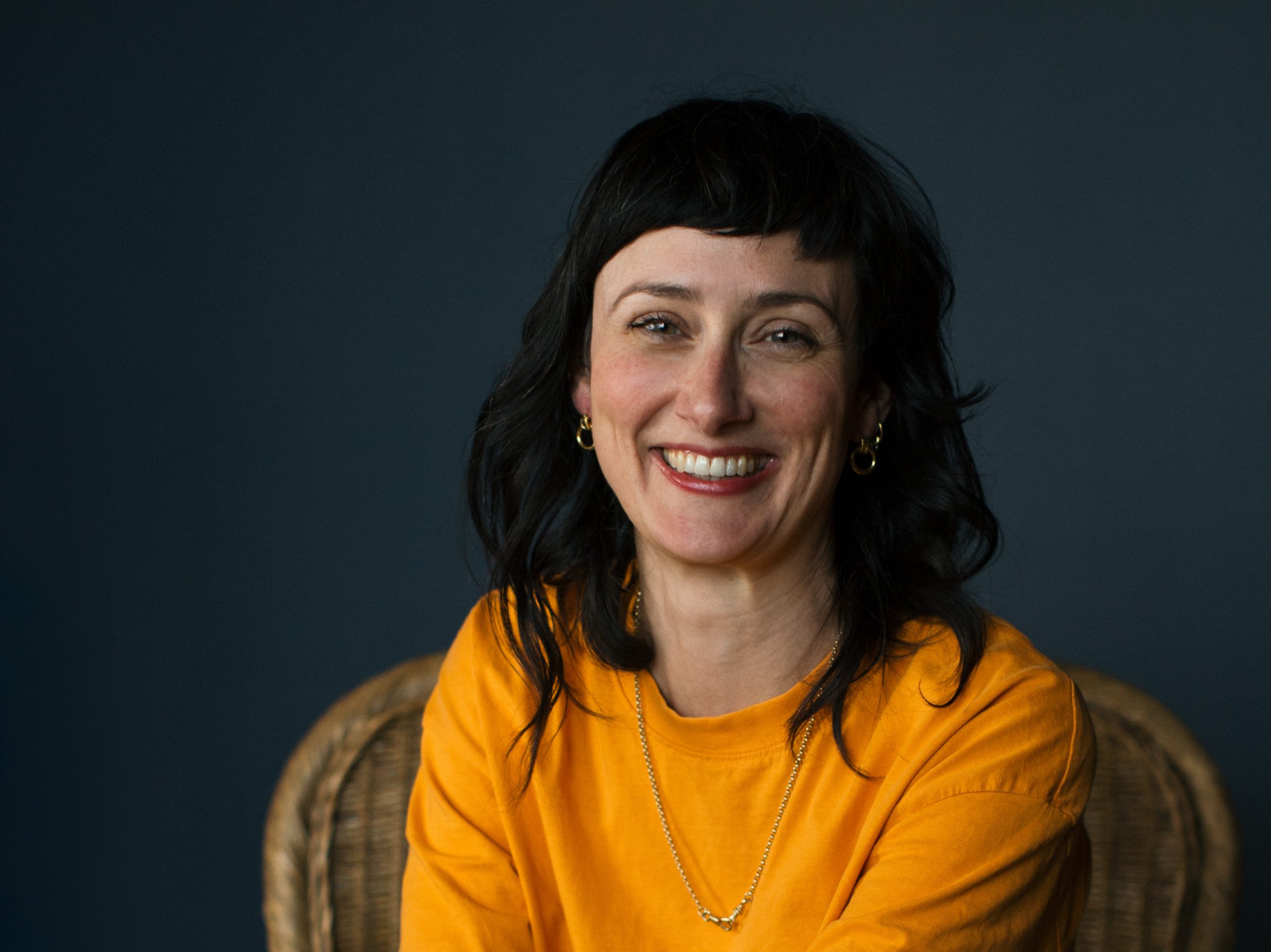 Sally is a slim, white, femme presenting person. She is sat, portrait style on a wicker chair, wearing a yellow t-shirt. Sally has long dark hair and is smiling brightly.