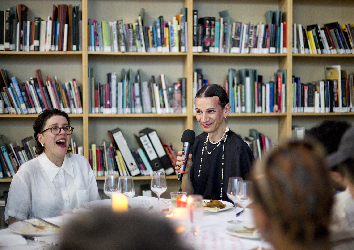 Person holding a microphone seated at the head of a dinner table. Bookshelves in the background.