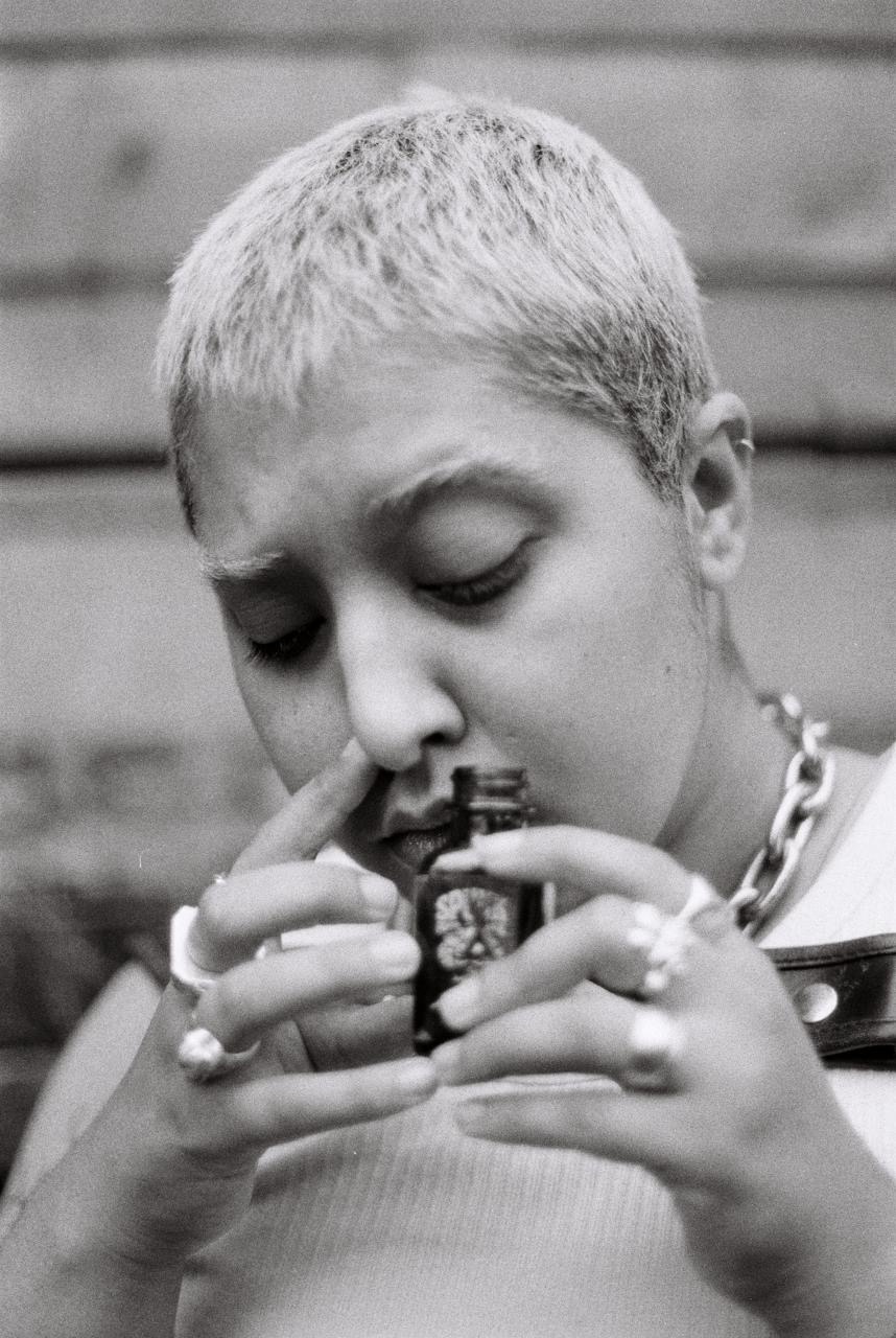 A black and white photograph of an Iranian trans-masculine person wearing a large chain necklace and harness, sniffing poppers.